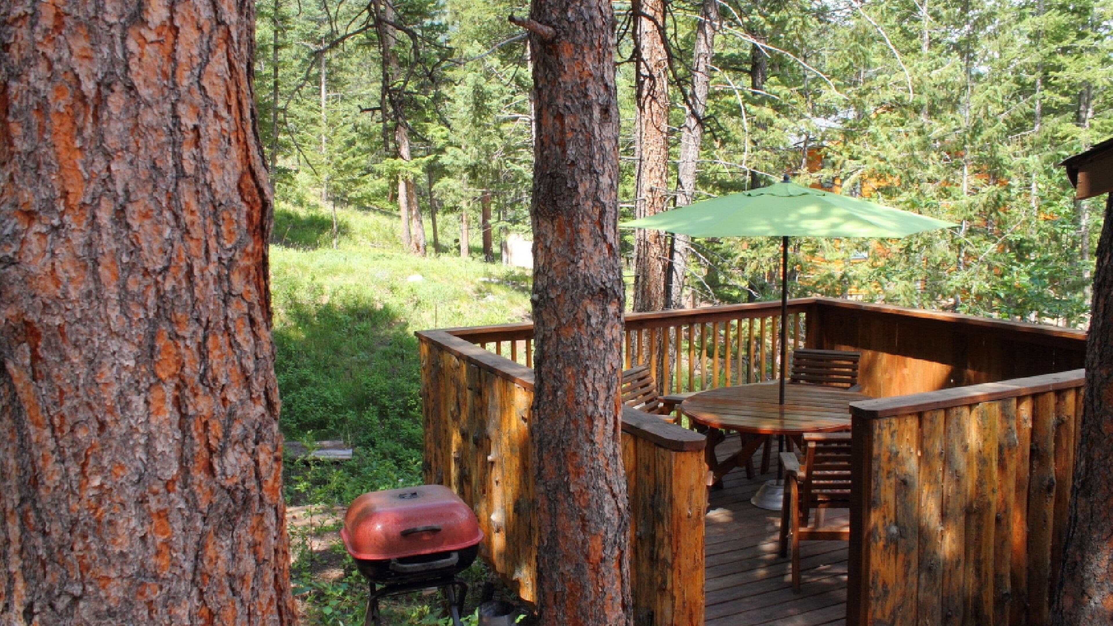 A wooden deck with a barbecue grill and a green umbrella, surrounded by trees.