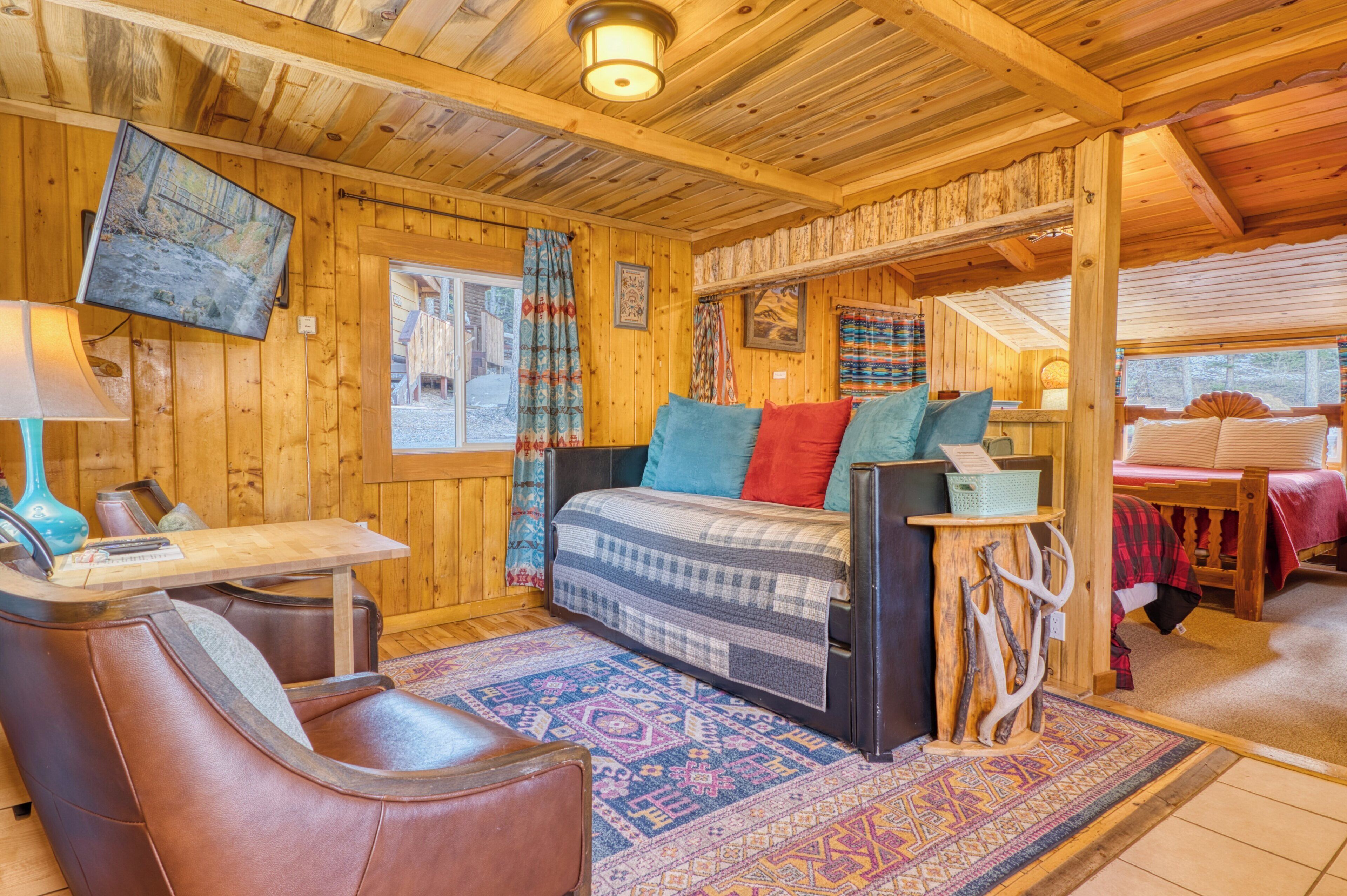A cozy cabin interior with a wooden ceiling, a bed with colorful pillows, a table with a lamp, and a mounted TV.
