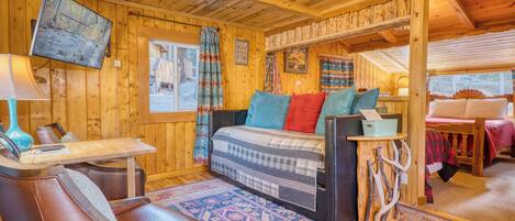 A cozy cabin interior with a wooden ceiling, a bed with colorful pillows, a table with a lamp, and a mounted TV.