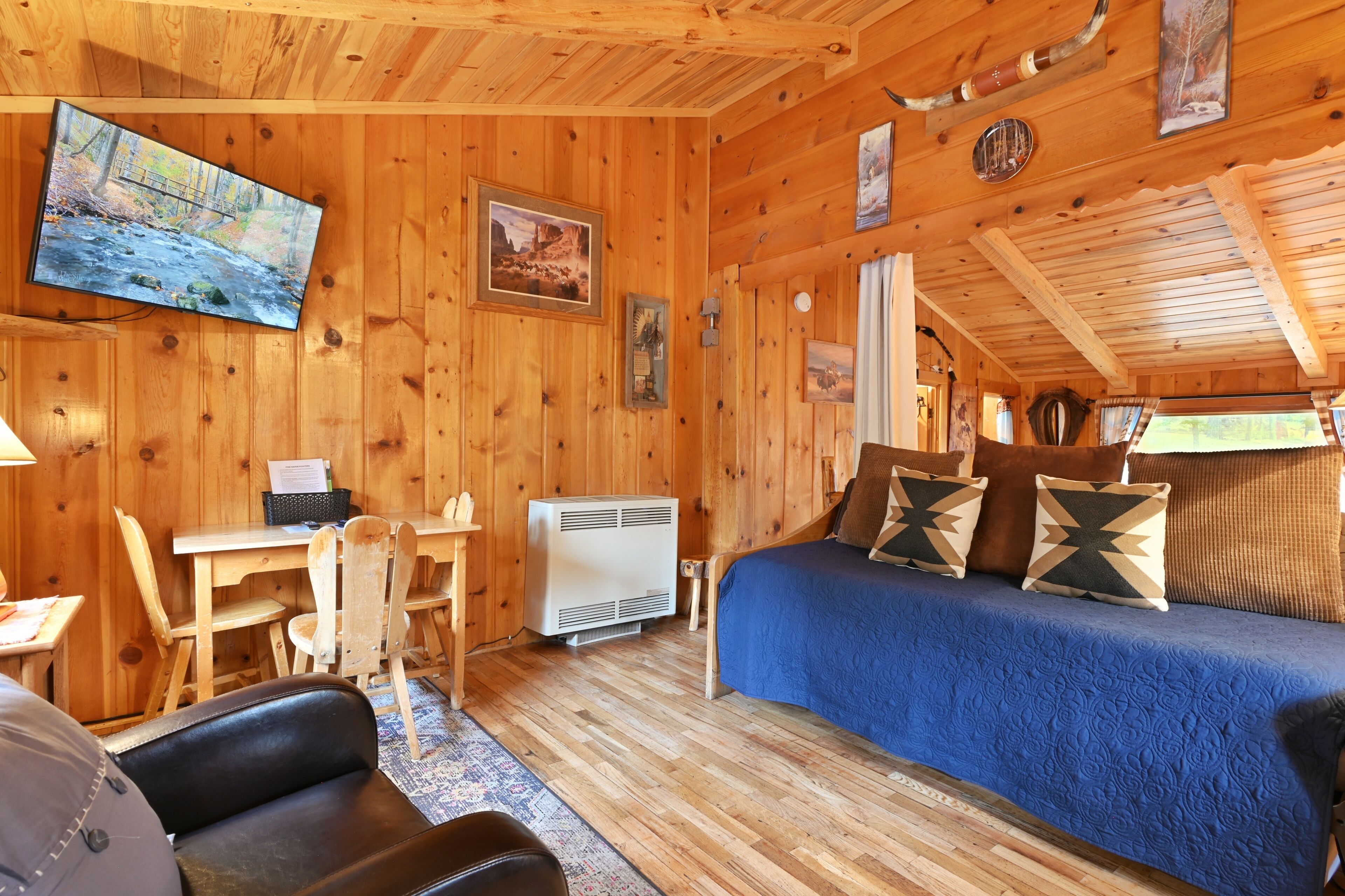 A cozy cabin interior with a wooden interior, a bed with blue bedding, a desk, and a television mounted on the wall.