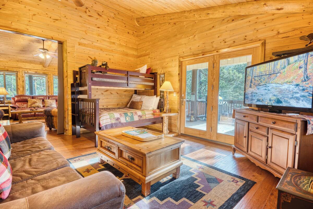 A cozy cabin interior with a wooden ceiling, a bunk bed, a television, and a large window offering a view of the outdoors.