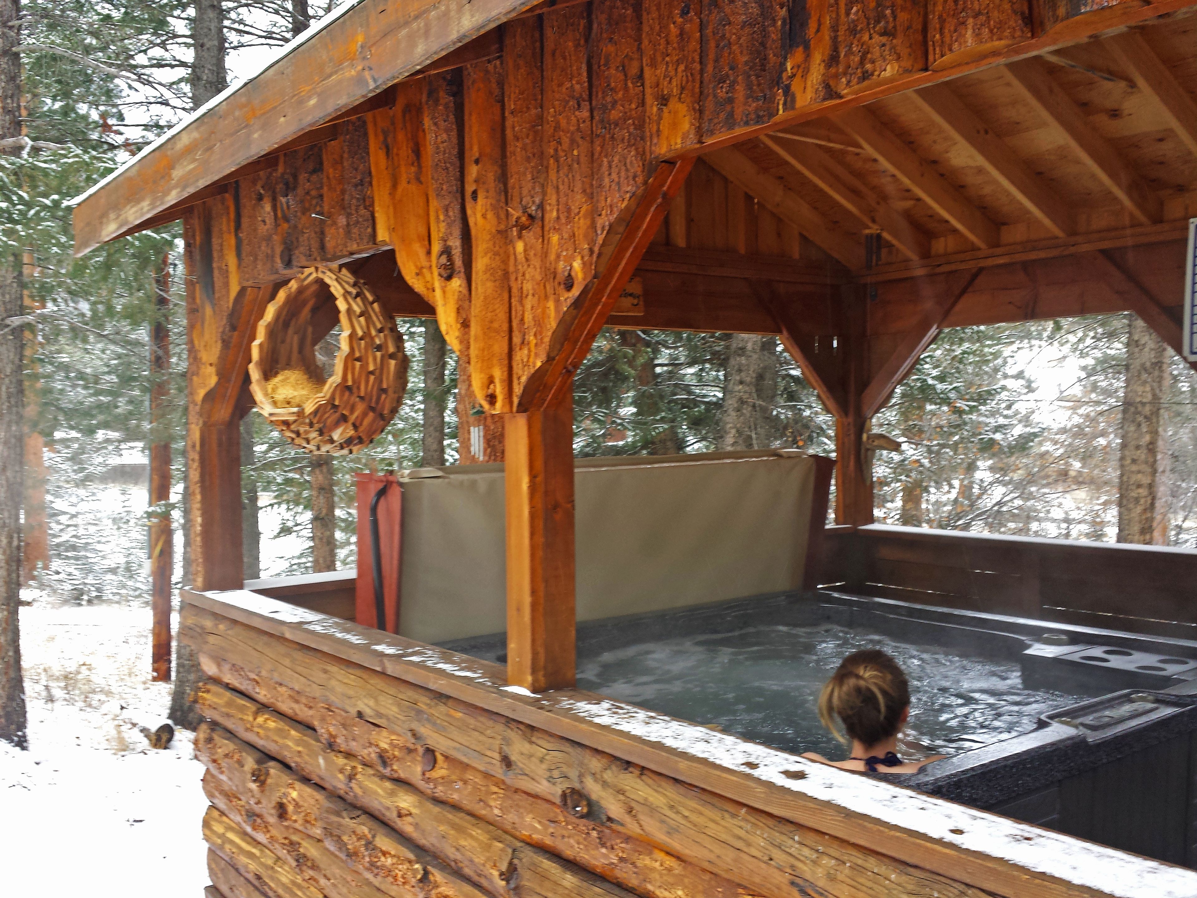 Una cabaña de madera con hidromasaje, rodeada de nieve y árboles.