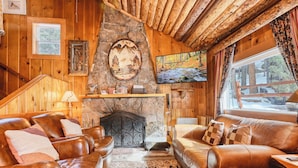 Interior acogedor de una cabaña con sofás de cuero, chimenea de piedra, paredes de madera y un ventanal con vistas a un paisaje nevado.