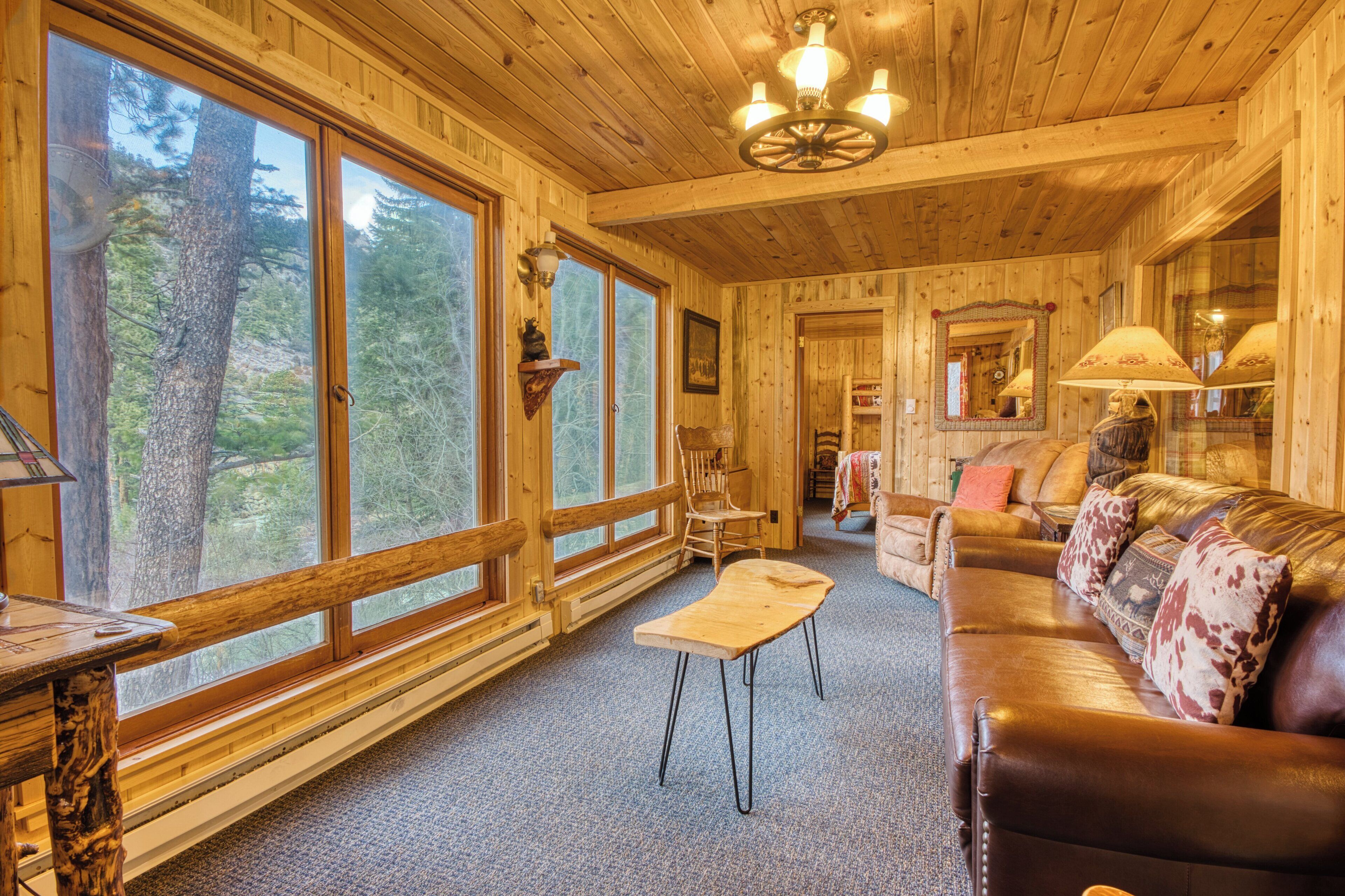 A wooden cabin interior with large windows, a leather sofa, a wooden bench, and a chandelier.