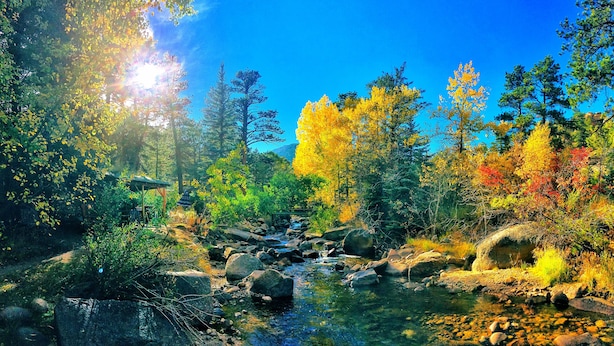 Paisaje natural con un arroyo, rocas y árboles en diversos tonos otoñales.