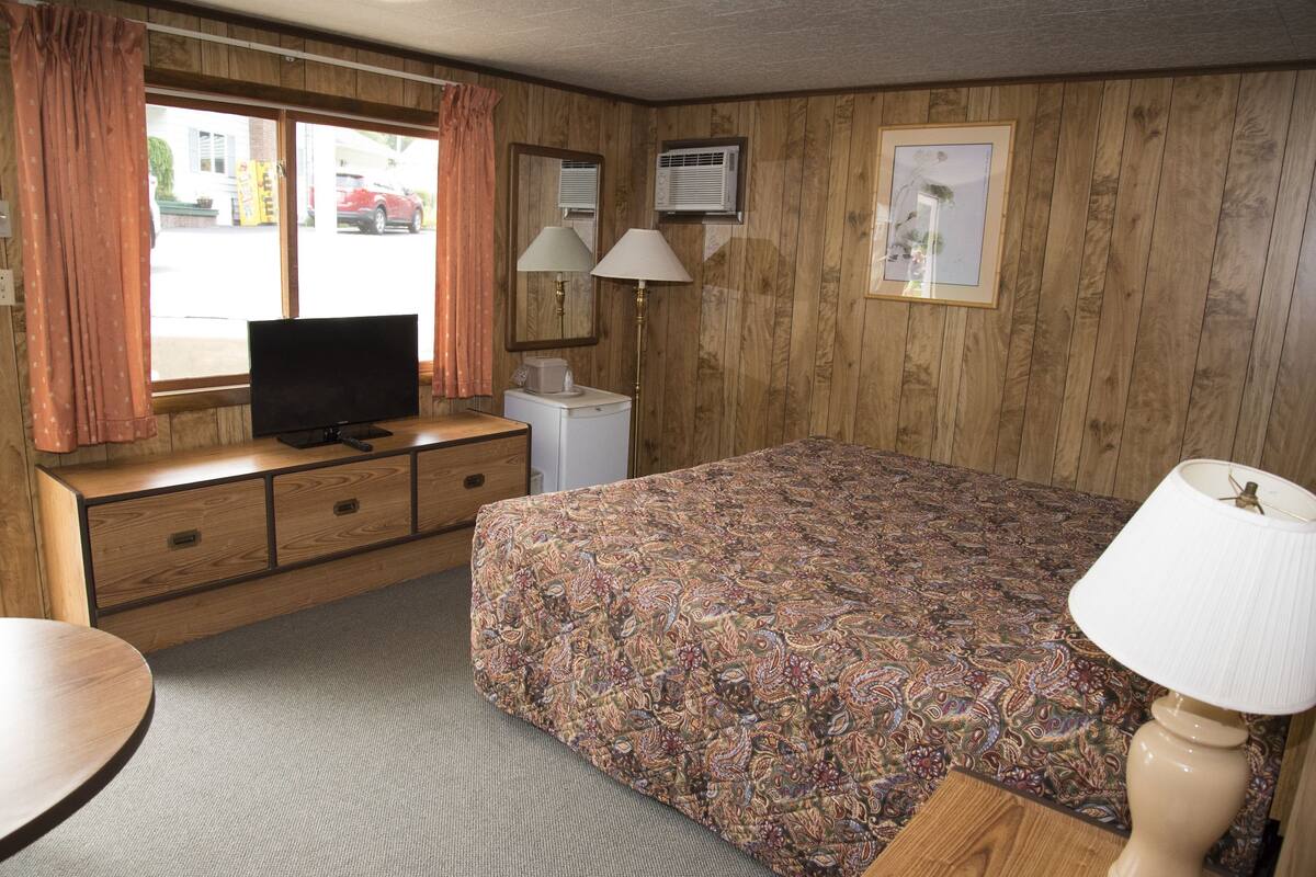 A motel room with a bed, a television on a wooden dresser, a desk with a lamp, and a window with curtains.