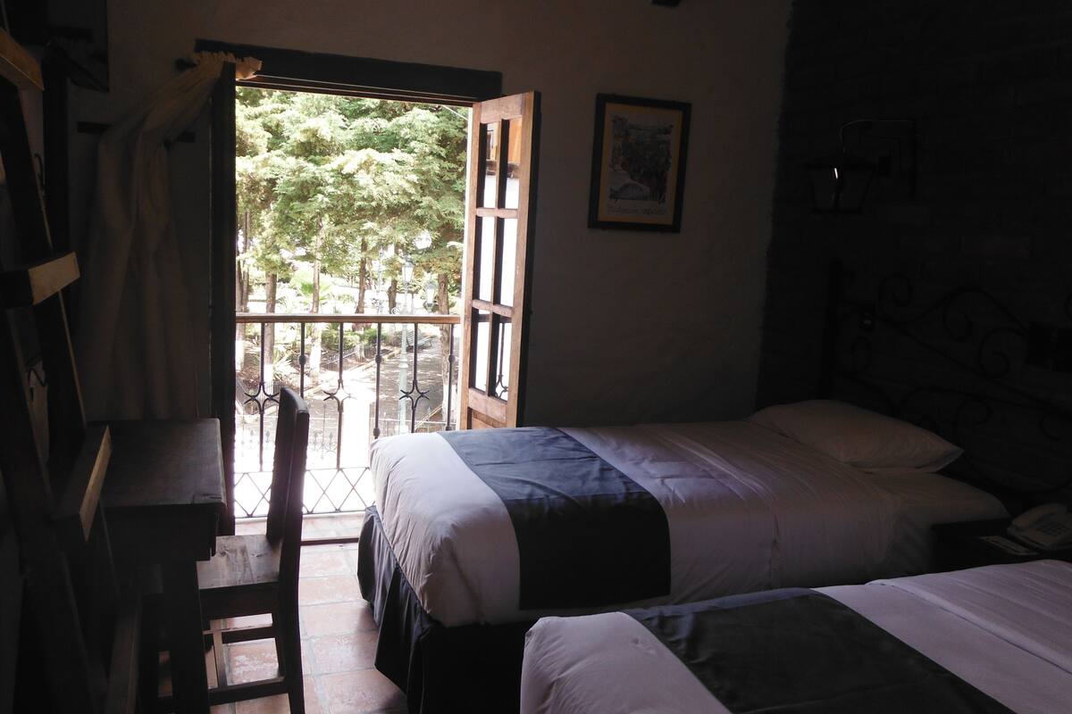 Une chambre avec deux lits, un bureau et une chaise. Elle dispose d’un balcon avec vue sur les arbres et une rambarde.