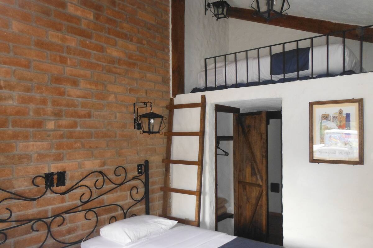 Une pièce avec un mur en briques, un cadre de lit en métal, une échelle en bois et un tableau encadré accroché au mur.