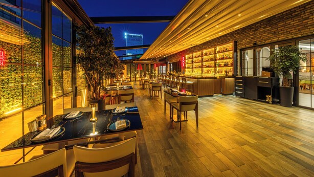 Un restaurante moderno con barra, suelo de madera y amplios ventanales con vistas a un paisaje urbano.