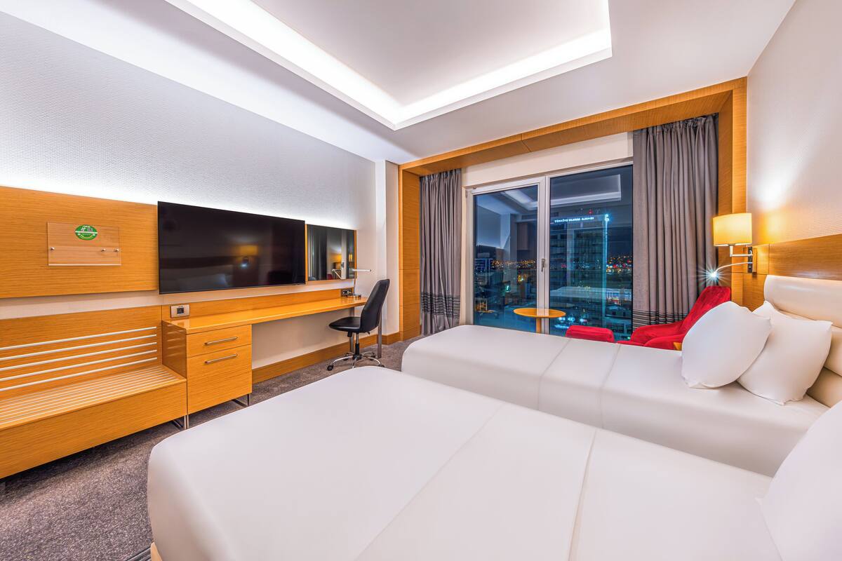 A hotel room with a bed, a desk with a TV, a chair, and a view of the city at night.