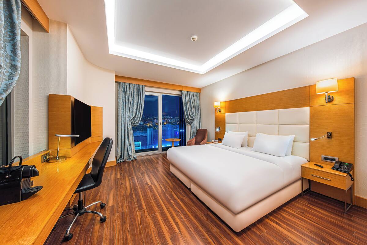 A modern hotel room with a large bed, a desk with a chair, a TV, and a city view through the window.
