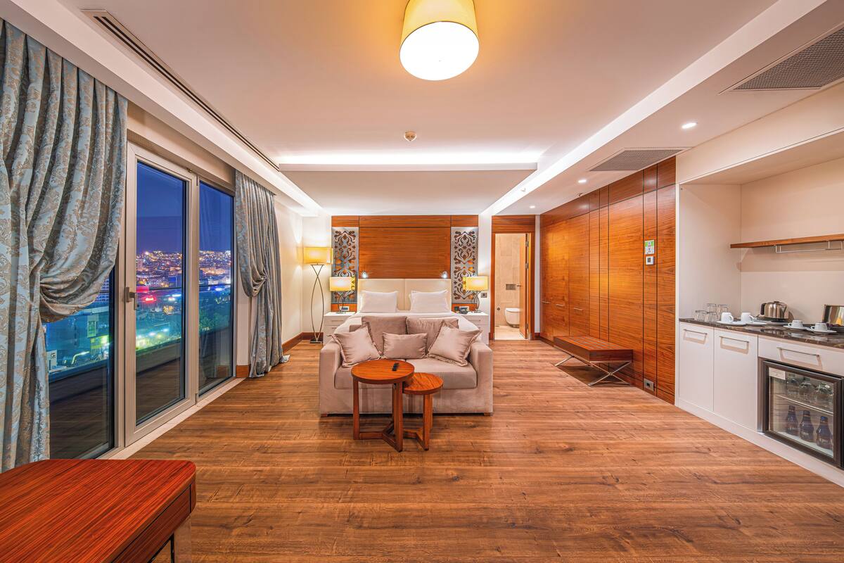 A modern hotel room with a sofa, a small table, a bench, a kitchen area, and a view of the city at night.