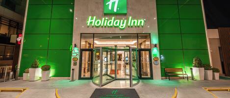 The exterior of a Holiday Inn with a prominent green logo, a glass entrance, and a well-lit facade.