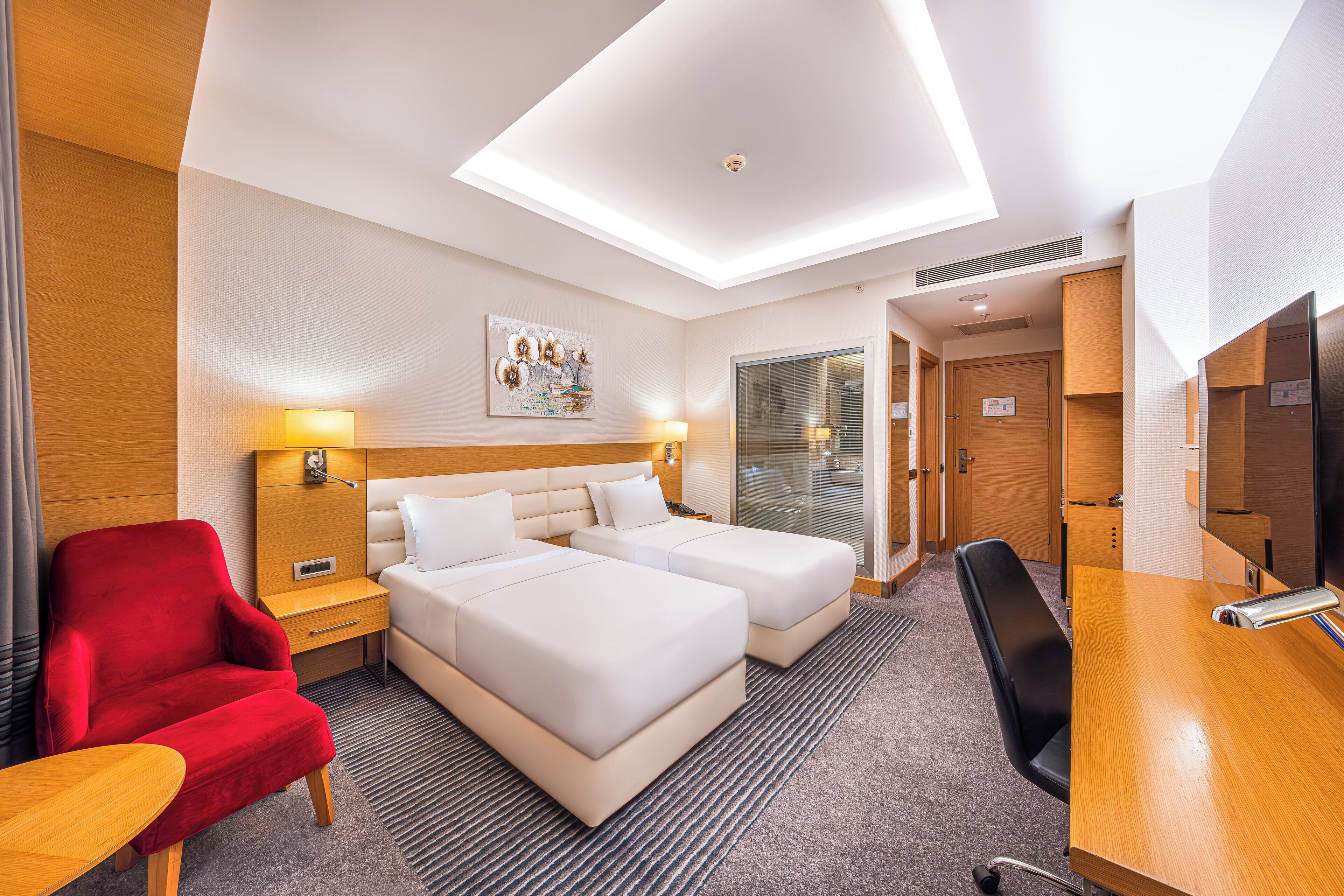 Ein Hotelzimmer mit zwei Betten, einem roten Sessel, einem Schreibtisch und einem großen Fenster.