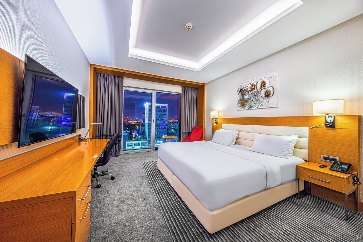 A hotel room with a large bed, a desk with a TV, a nightstand with a phone, and a view of the city at night.