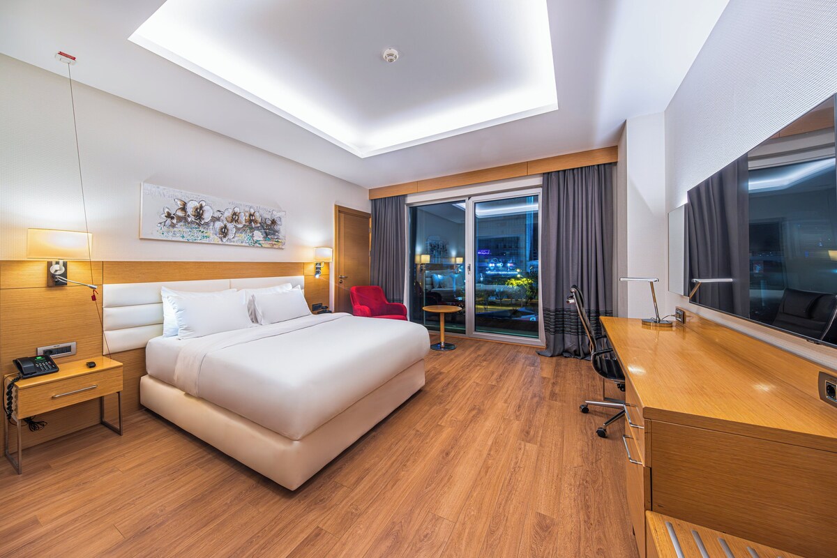 A hotel room with a large bed, a desk, a chair, a small table, and a view of the city at night.
