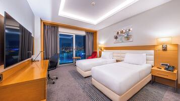 A hotel room with a desk, two beds, a city view, a painting, and a nightstand with a phone.