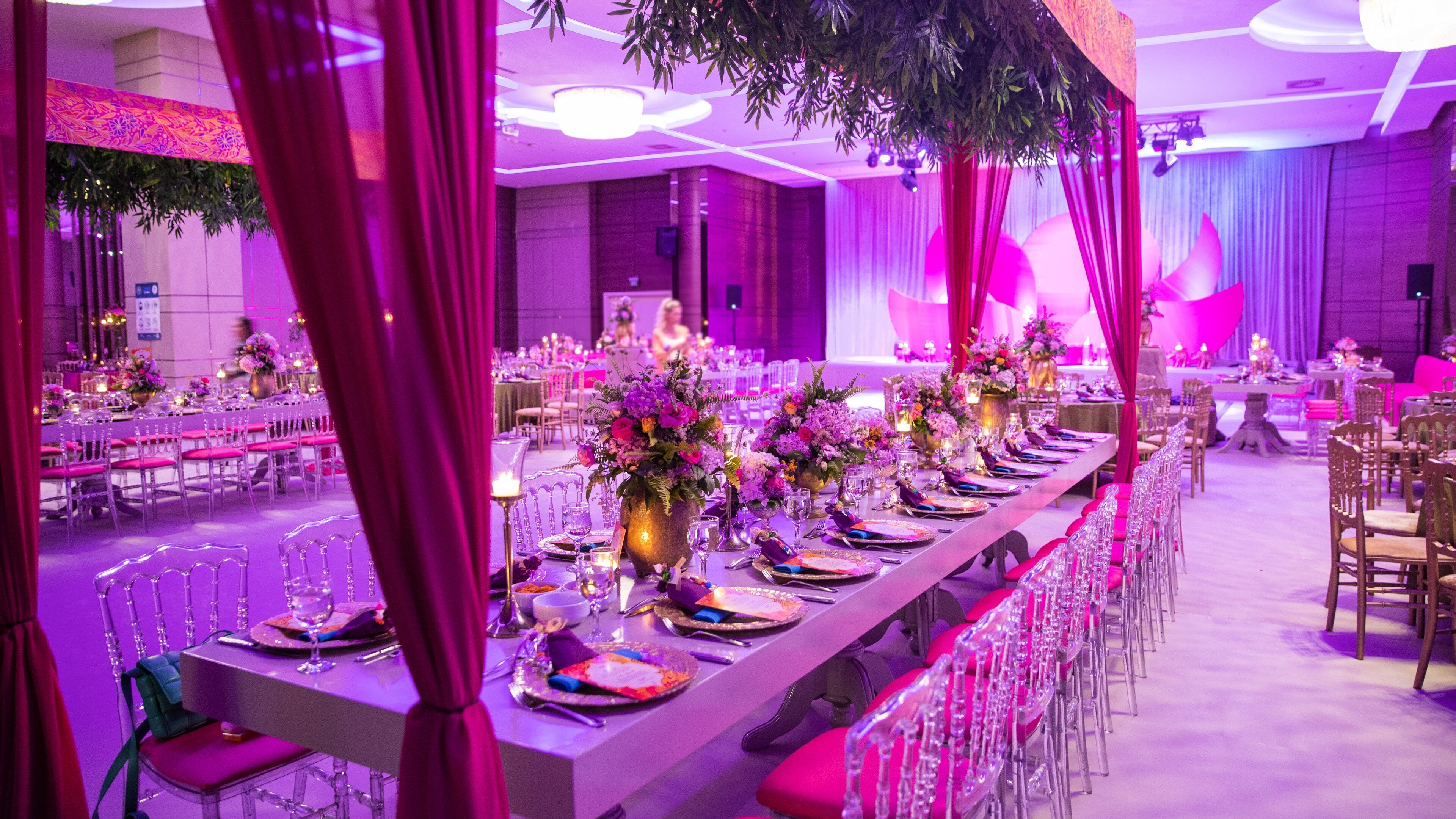 Una larga mesa de banquete con centros de flores, sillas rosas y una sala iluminada en morado.