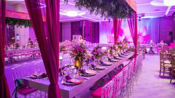 Una larga mesa de banquete con centros de flores, sillas rosas y una sala iluminada en morado.
