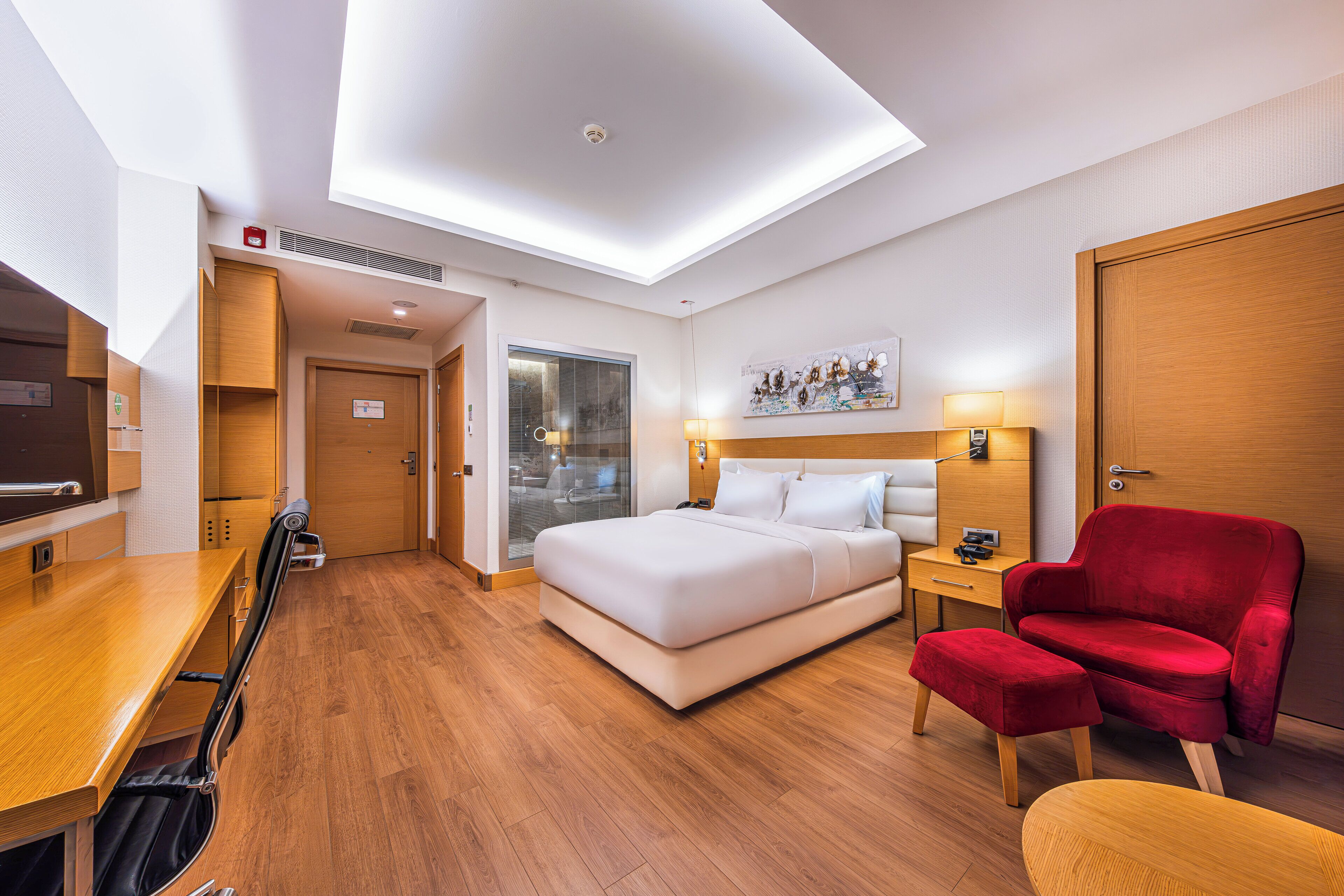 A hotel room with a bed, a desk, a red chair, a wooden floor, and a ceiling light.