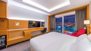 Habitación de hotel con una cama grande, un escritorio con televisor, minibar y vistas a la ciudad.