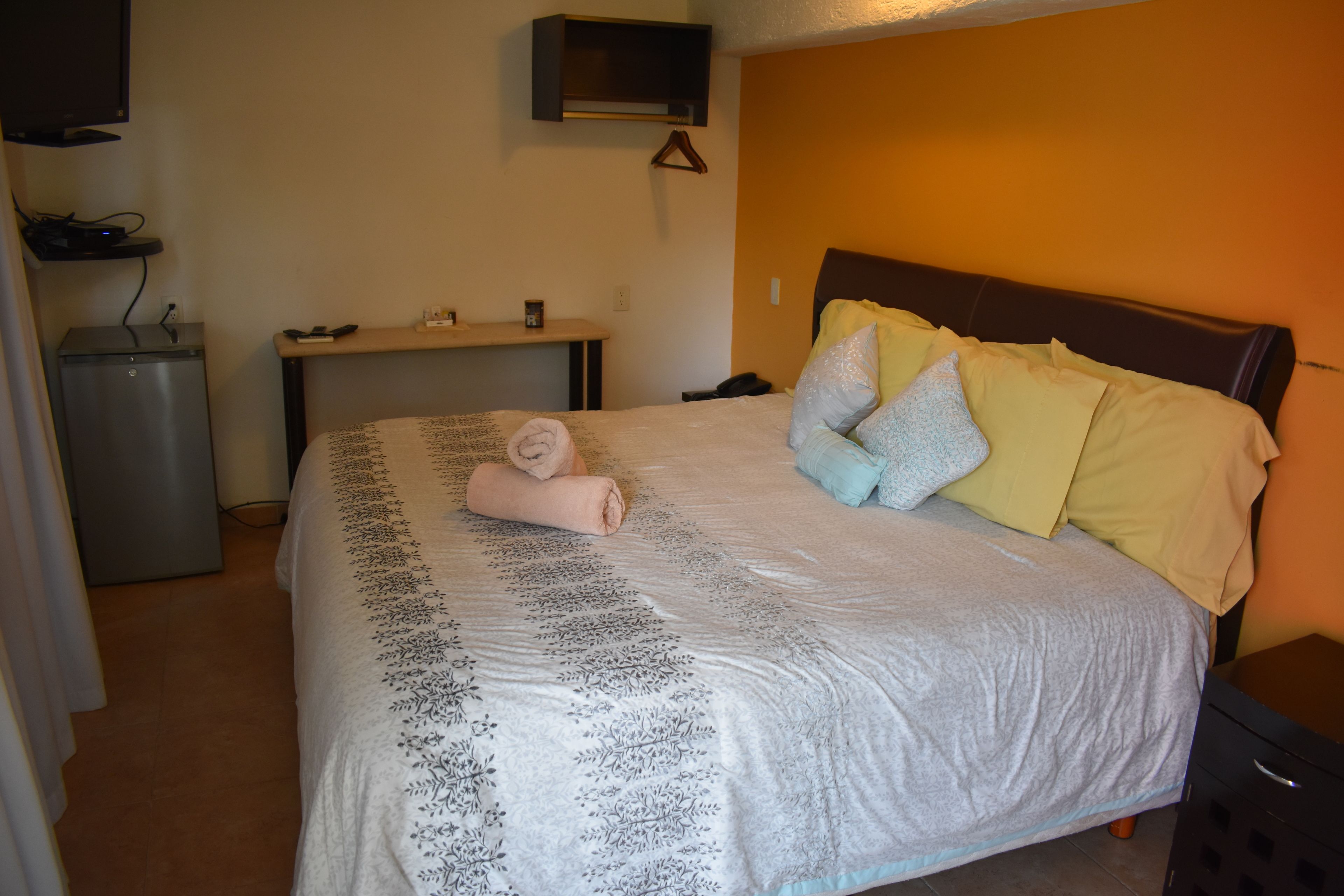 Un lit bien fait, recouvert d’une couvre-lit à motifs, une table de chevet avec une lampe, et un petit bureau avec un ordinateur et un téléviseur fixé au mur.