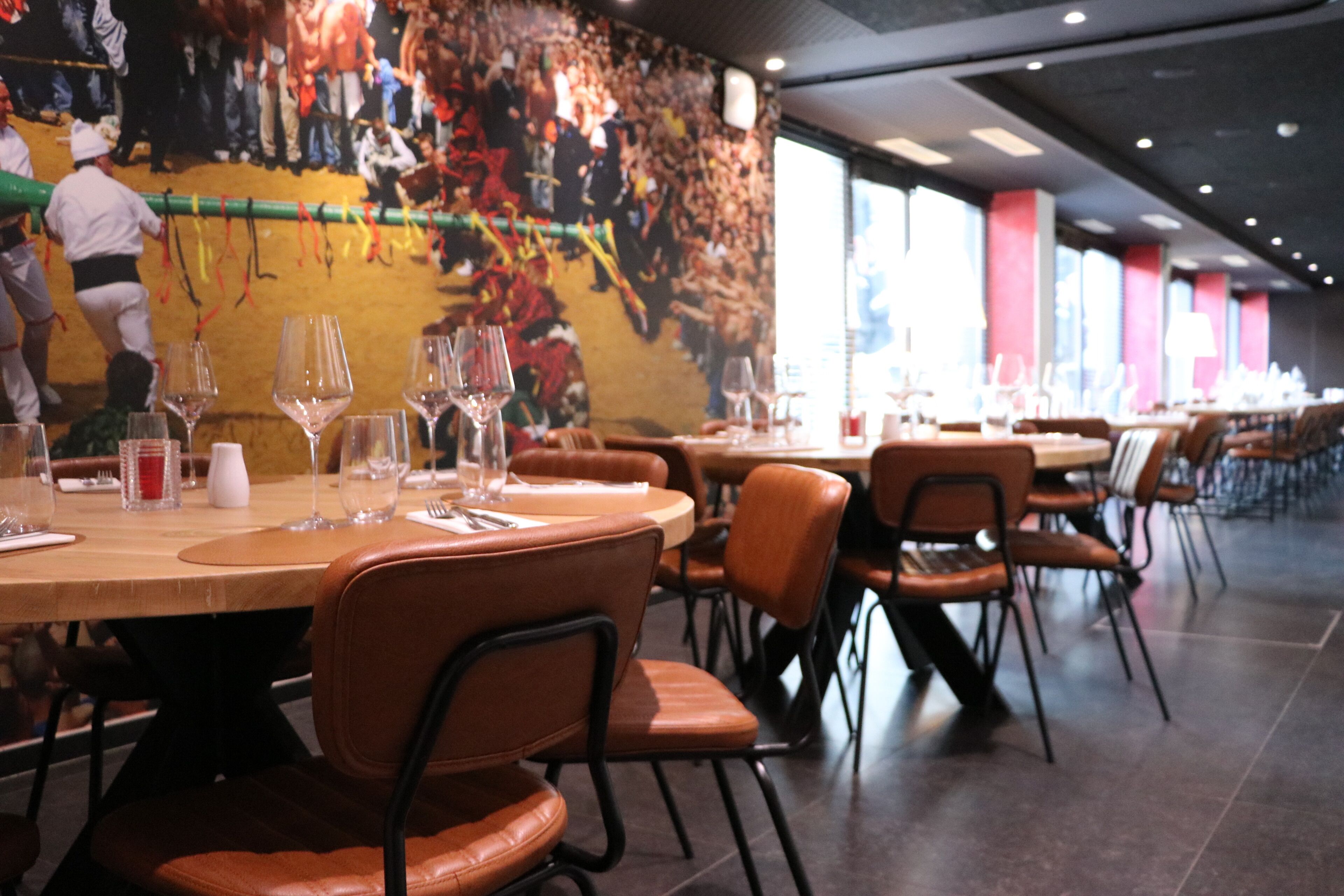 Un restaurante con mesas redondas y sillas de cuero, ventanas amplias y un mural en la pared.