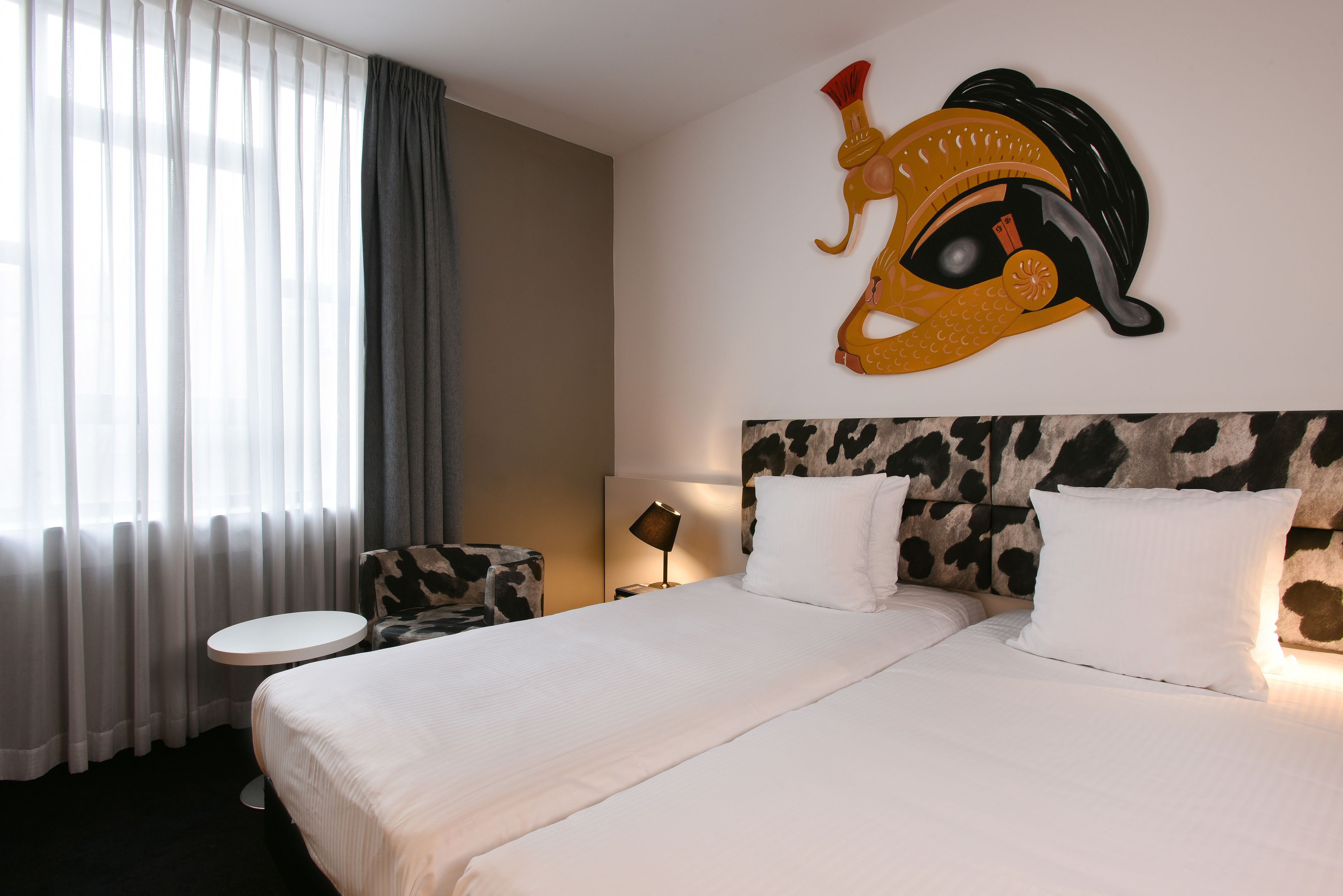 Una habitación de hotel con una cama grande, una mesita redonda, una lámpara y una decoración de pared original.