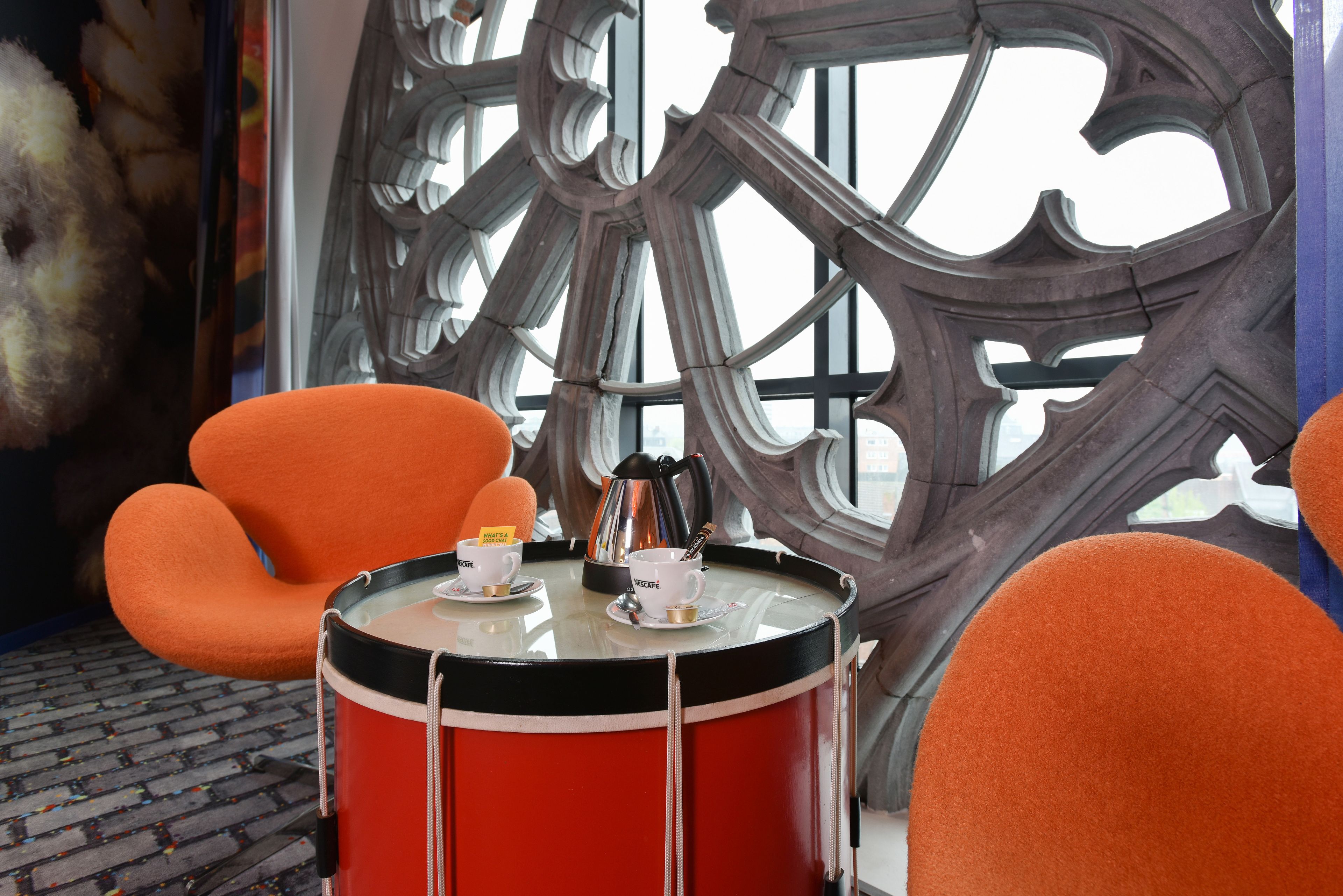 Een kamer met een groot, versierd raam, een rode trommel en oranje stoelen.