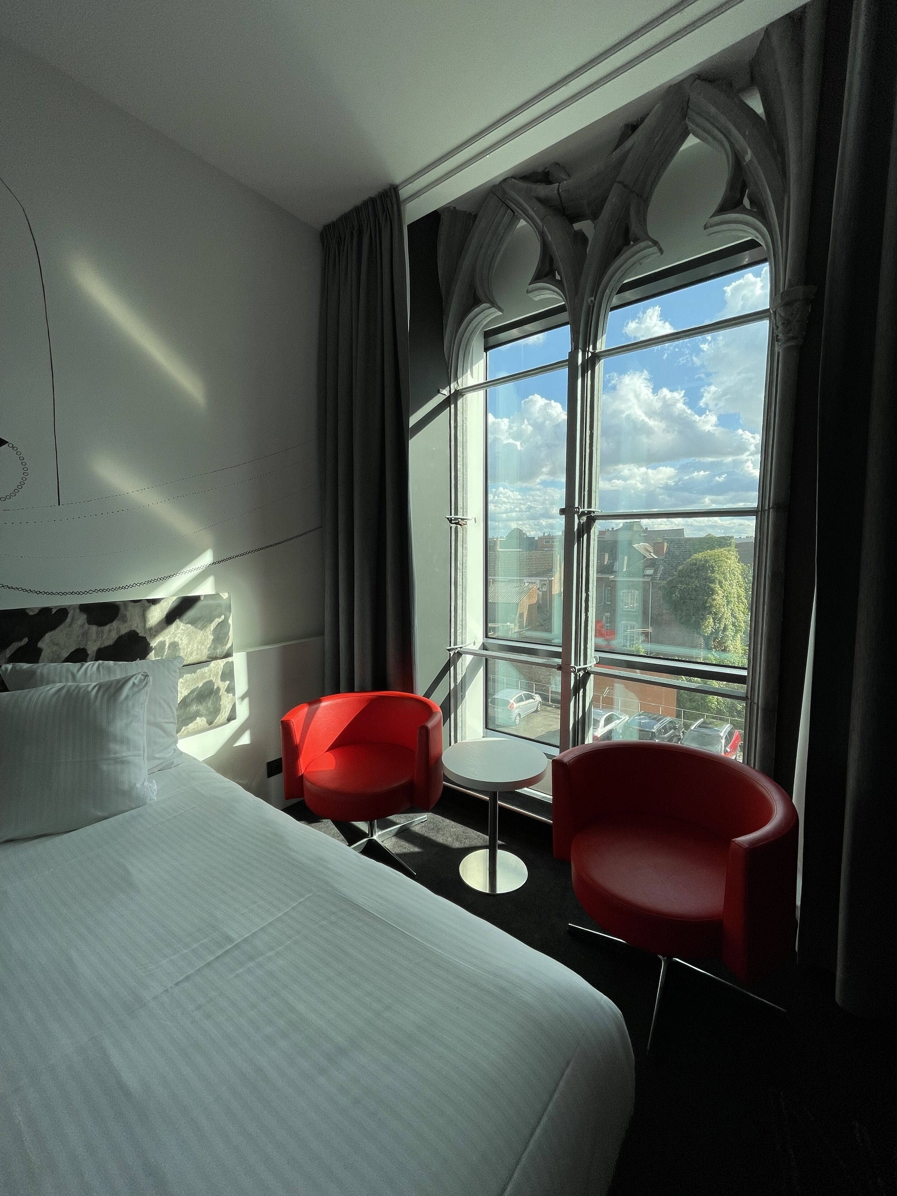 A hotel room with a large bed, a small table, two red chairs, and a large window with a view of the city.