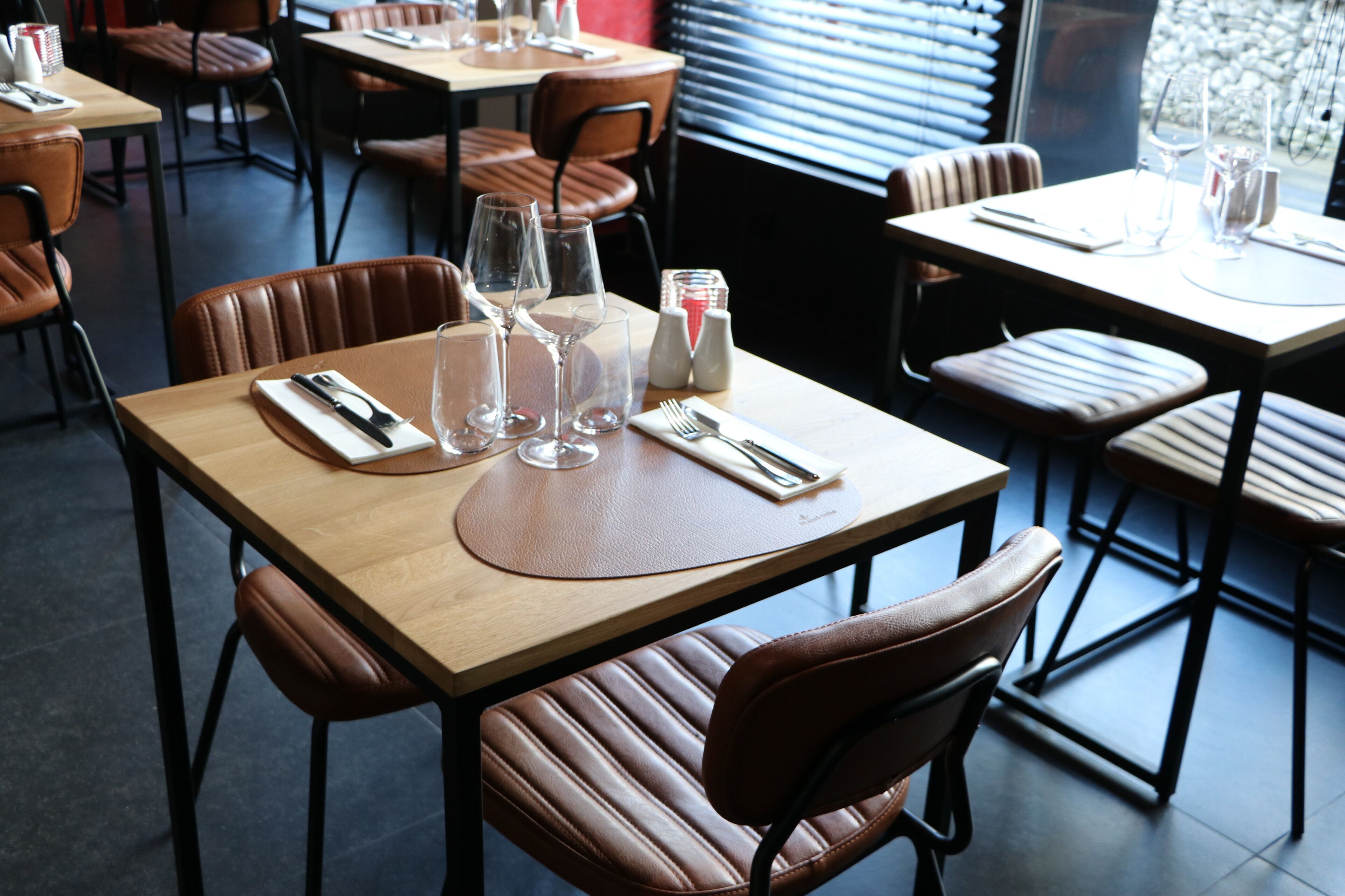 Interior de restaurante con mesas de madera, sillas de cuero marrón y cristalería transparente en cada mesa.
