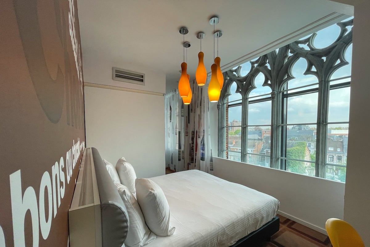 A bedroom with a large bed, a window with a city view, and a unique architectural window design.