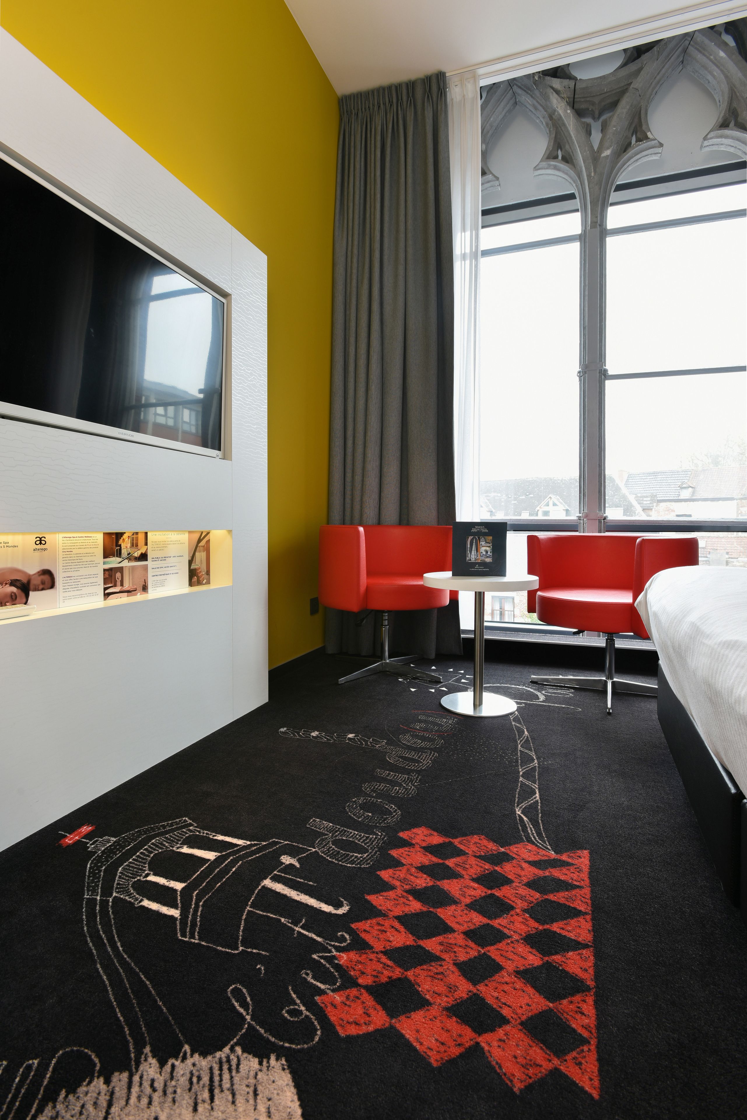 A hotel room with a bed, a TV, a red chair, a small table, and a window with a view of buildings.