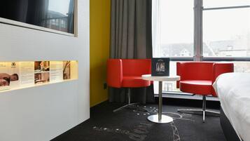 Une chambre d’hôtel avec un lit, une télévision, un fauteuil rouge, une petite table et une fenêtre donnant sur des immeubles.