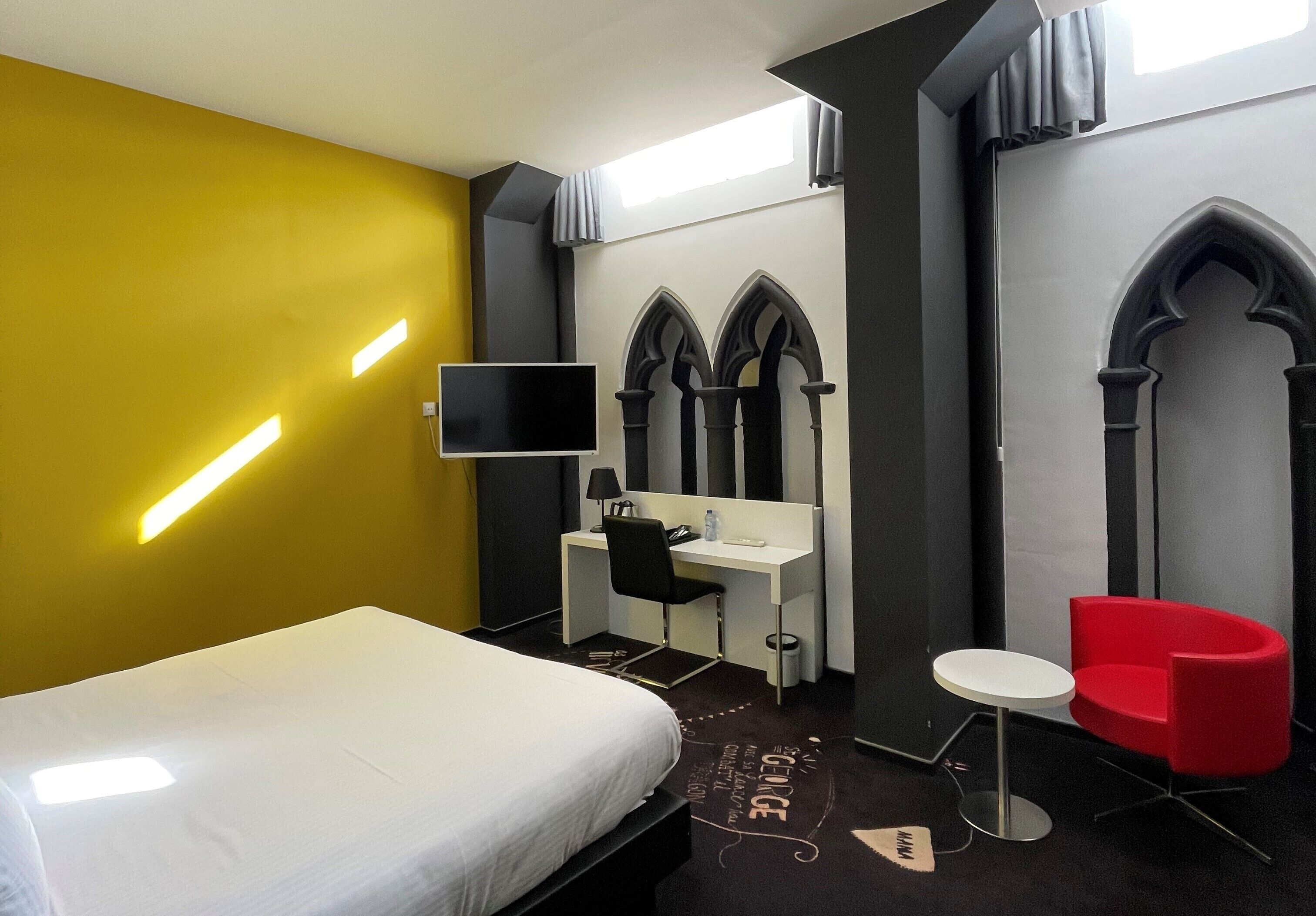 A hotel room with a bed, a desk, a chair, a TV, and a red chair.
