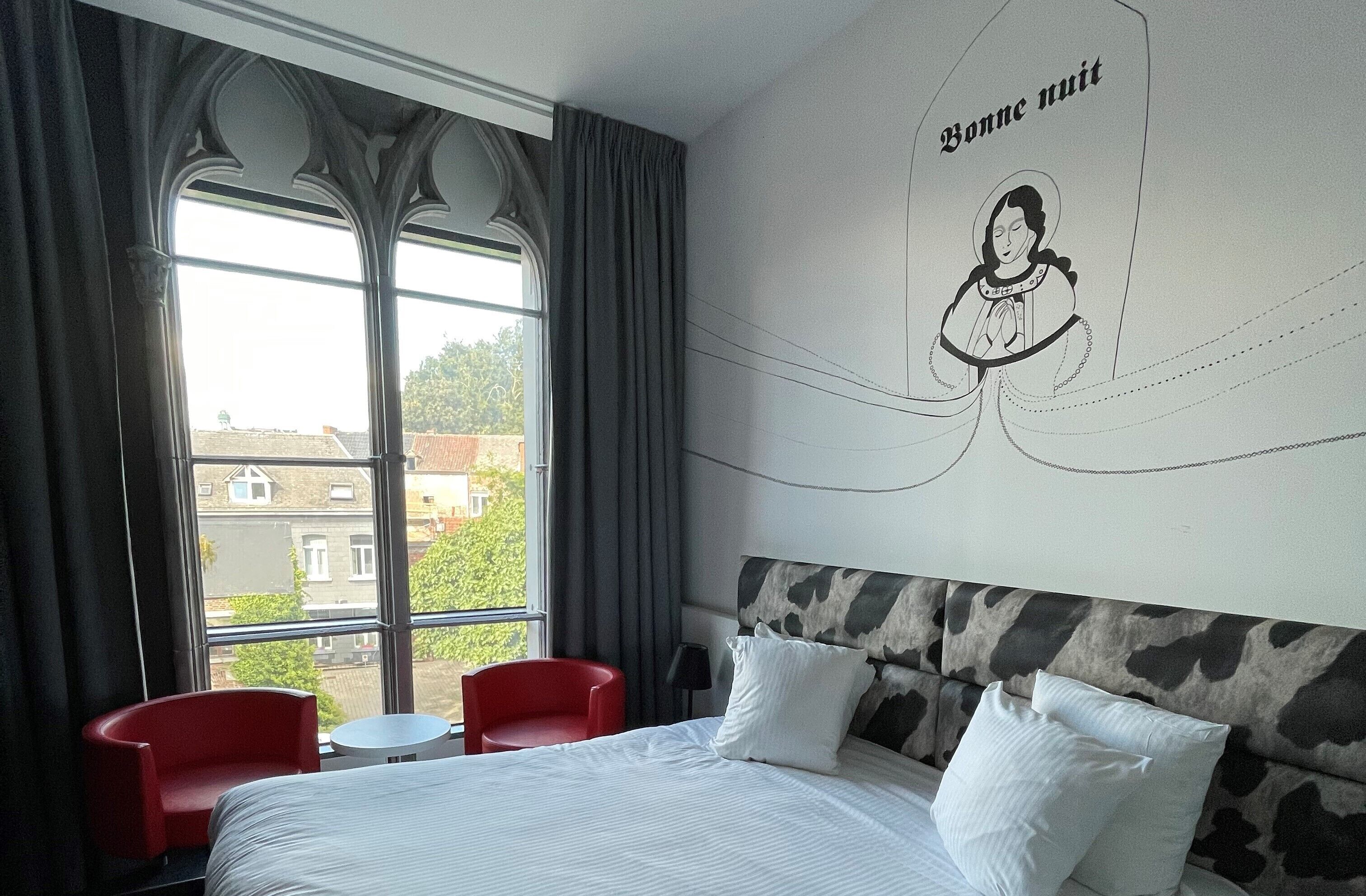 A hotel room with a large bed, two red chairs, a small table, and a view of buildings through large windows.