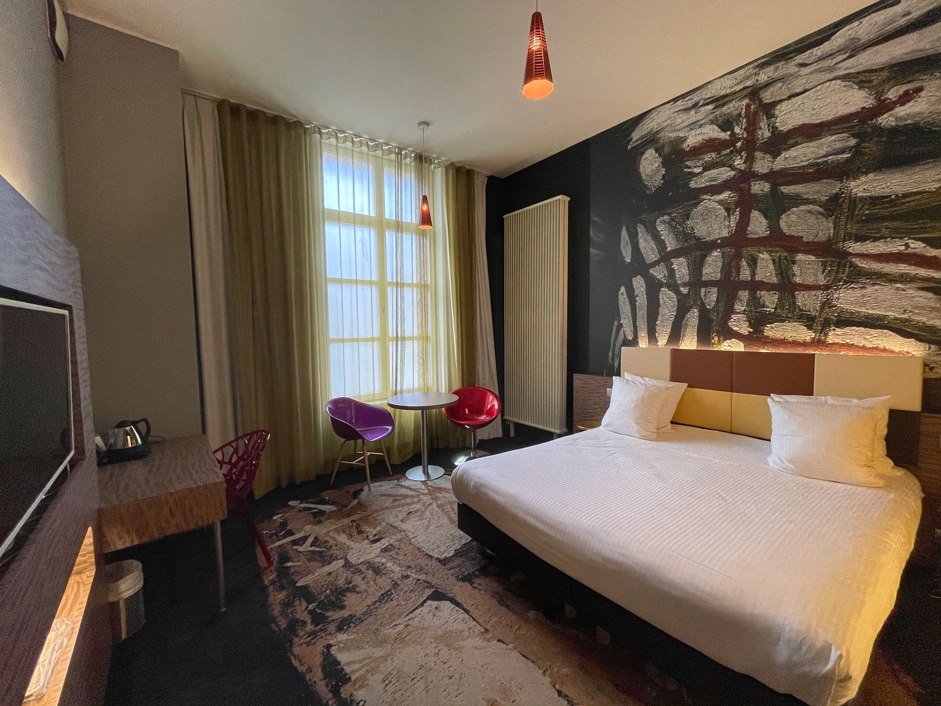 Une chambre d’hôtel avec un grand lit, un bureau, une chaise, une petite table et un tableau au mur.