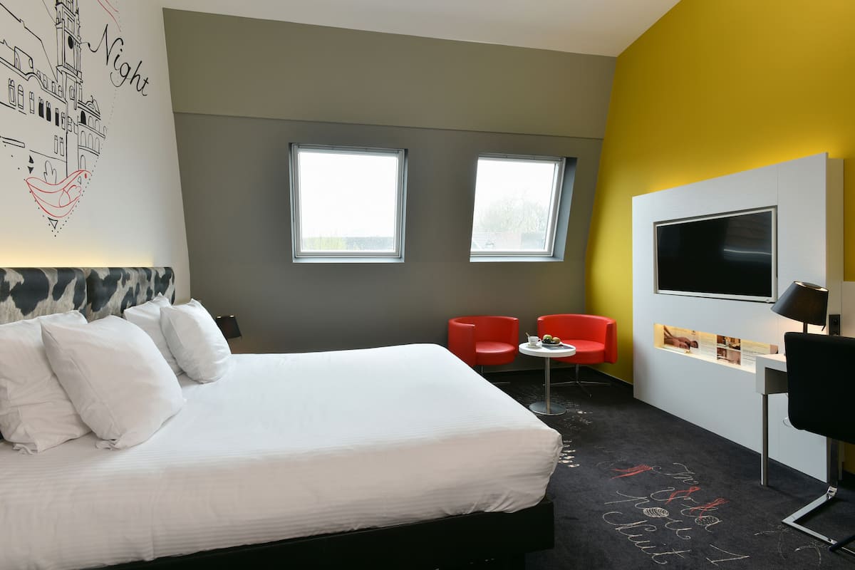 A hotel room with a bed, two windows, a desk with a TV, and red chairs.