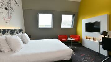 Een hotelkamer met een bed, twee ramen, een bureau met een televisie en rode stoelen.