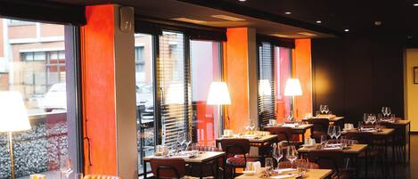 Un restaurante con mesas y sillas, iluminado por lámparas y grandes ventanales.