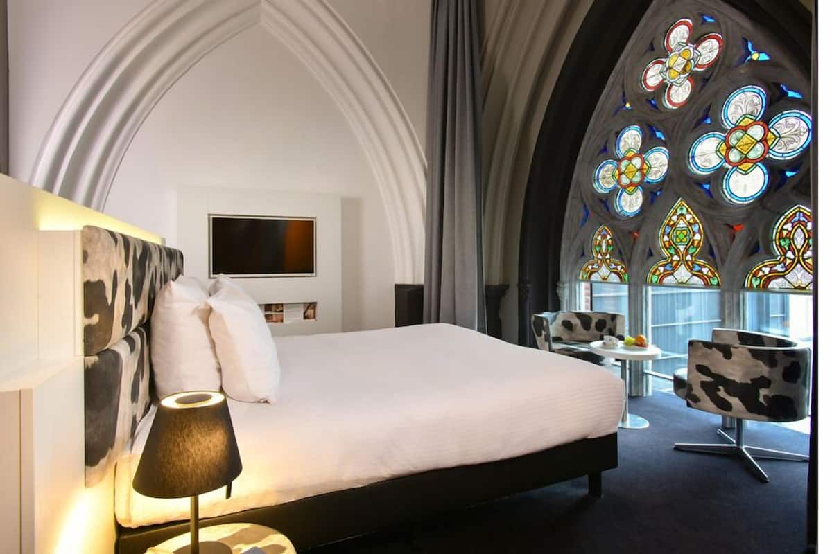 A hotel room with a large bed, a TV, a modern chair, and a stained glass window.
