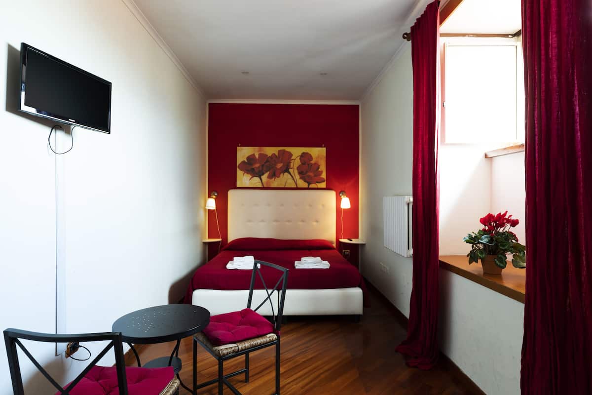 A hotel room with a large bed, a television mounted on the wall, a small table with a chair, and a window with red curtains.