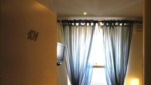 Une chambre d’hôtel avec un plancher en bois, un lit, une chaise, un bureau avec une lampe et une fenêtre avec des rideaux légers.