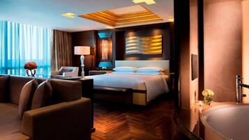 A hotel room with a large bed, a sitting area with a sofa and armchair, a desk with a lamp, and a bathtub.