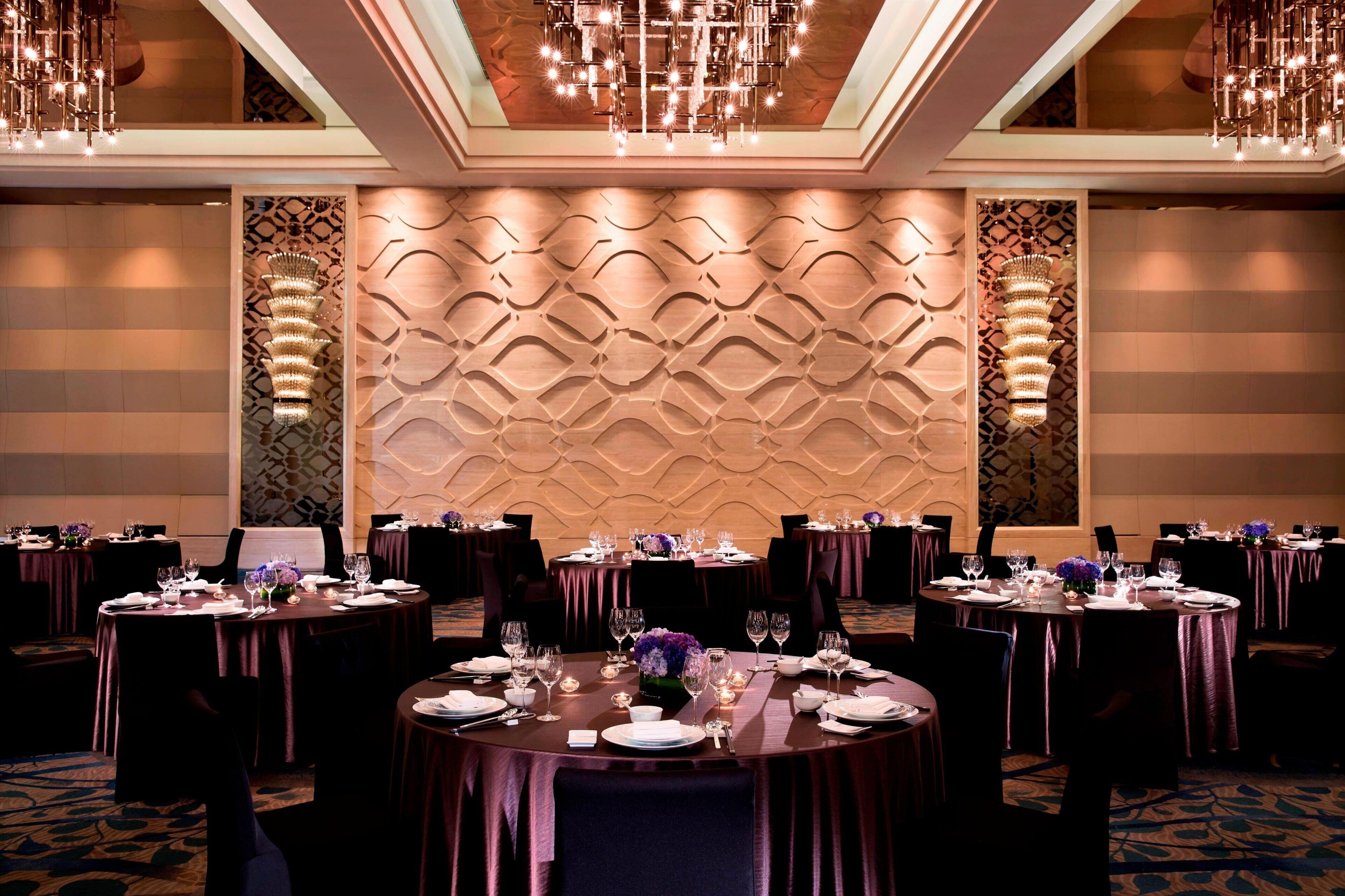 A banquet hall with round tables set for dining, featuring a patterned wall and chandeliers.