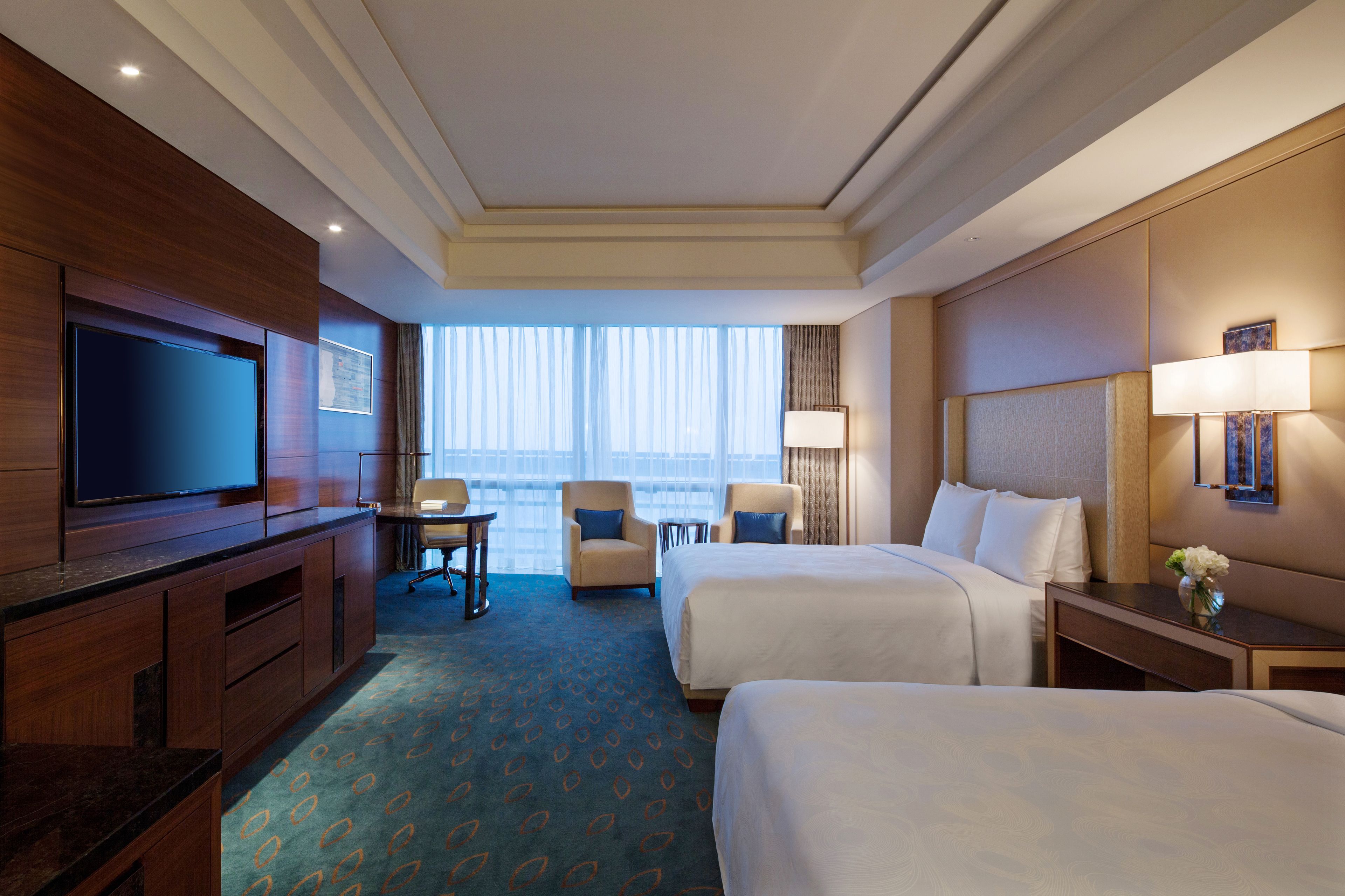 A hotel room with two beds, a TV, a desk, and a window with curtains.