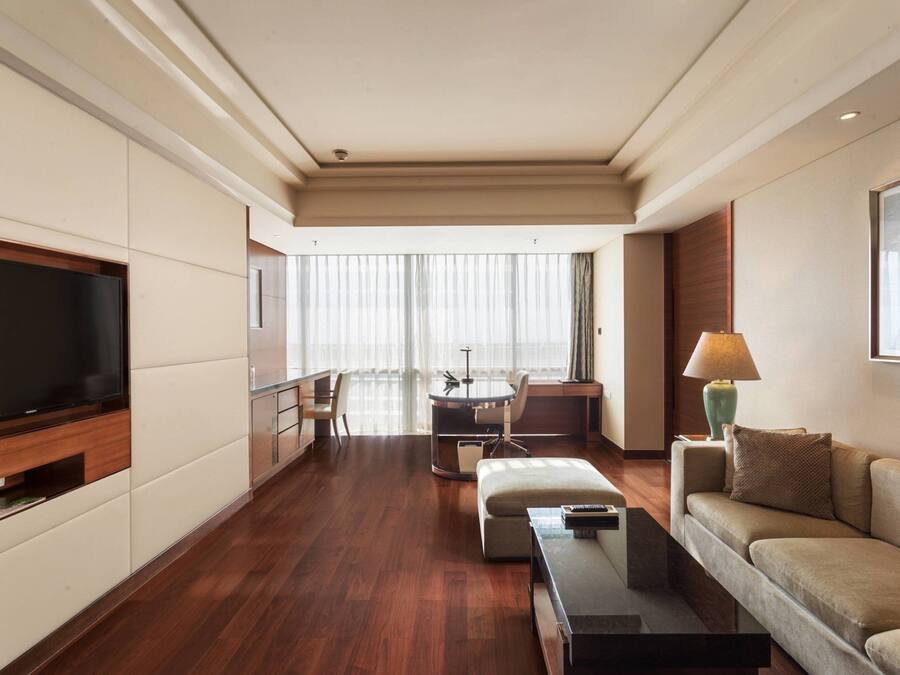 Una habitación de hotel moderna con televisor de pantalla plana, un sofá, una mesa de centro, una zona de comedor con mesa y sillas, y un ventanal con cortinas.