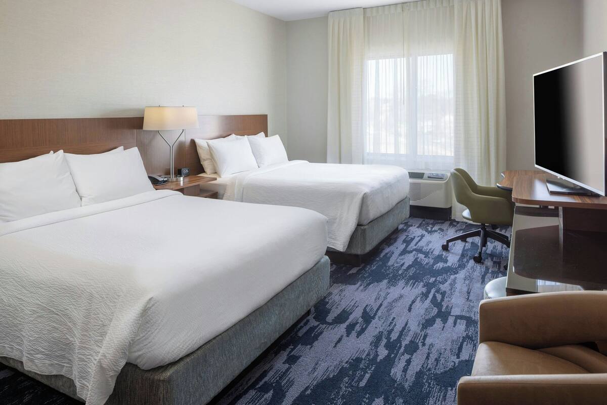 A hotel room with two beds, a desk, a chair, a TV, and a window with curtains.