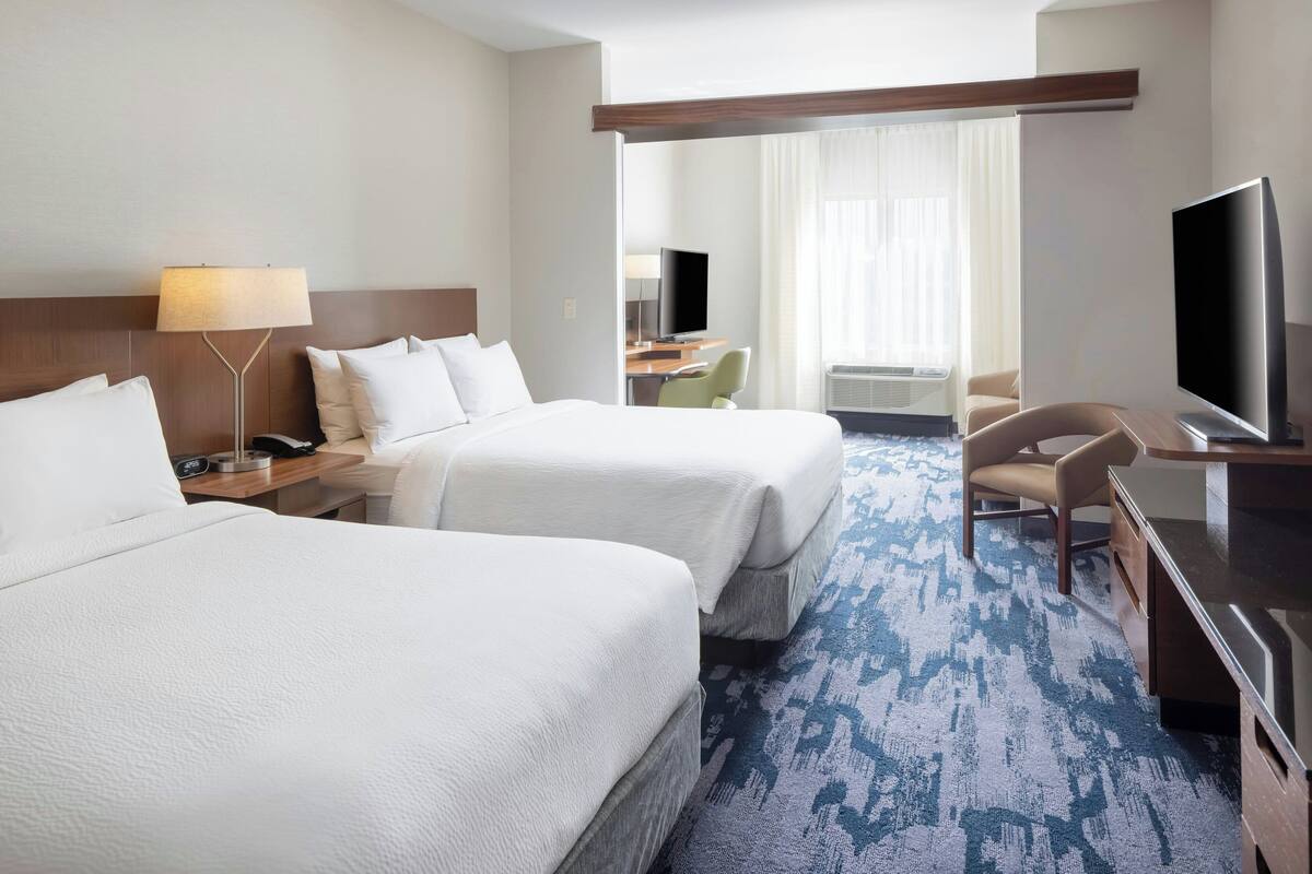 A hotel room with two beds, a desk with a chair, a television, and a large window with curtains.