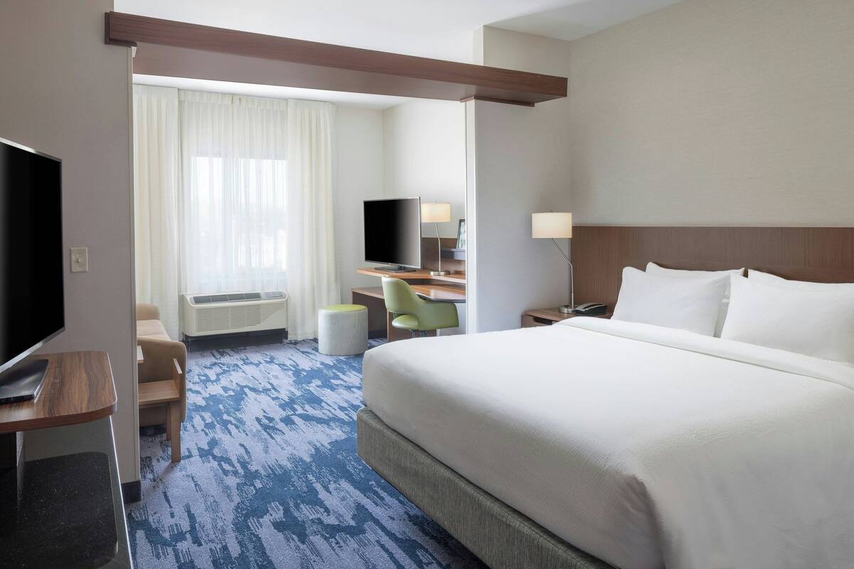 A hotel room with a large bed, a desk with a flat-screen TV, a chair, a lamp, and a window with curtains.